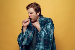 Young Sick Guy Wrapped in Checkered Plaid Coughing. Ill Handsome Person Feels Uncomfortably Unhealthy covering in Blue Wrap Isolated on Yellow Background. Concept of Cold