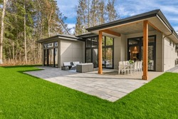 Exterior image of a contemporary home with flat roof and brown trim and lush green grass blue sky forest in the background furnished patio for outdoor dining and lounging