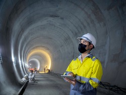 Young Asian tunnel engineering wearing high visibility jacket, face mask and white safety helmet working and using digital tablet in dark railway tunnel construction site area with copy space.