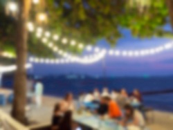 Abstract blurred of restuarant on the beach background with hanging light bulb bokeh on tree in the night party with many people, yellow string lights with bokeh decor in outdoor restaurant.