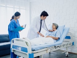 Asian elderly female patient dressed in light blue in bed and male doctor in white suit uses stethoscope to listen to heartbeat for check-up while assistant to record the symptoms for follow up.