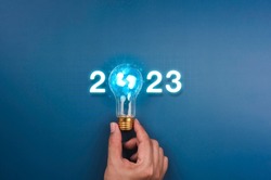 Shining 2023 calendar year numbers, neon style with creative trend light bulb holding by businessman's hand on digital network and blue background. Happy 2022 New Year greeting card with light bulb.