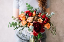 Very nice young woman holding a beautiful flower bouquet of fresh roses, peony, eucalyptus  in pastel cream, orange and vivid red colors on the grey background