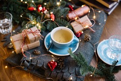 Delicious freshly brewed thyme tea in a blue cup standing on the warm cozy piled cover with red christmas ornamental hearts, little wrapped gifts, fireflies, pine cones on the wooden table background 