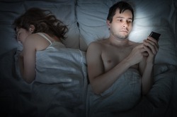 Young man is lying with girlfriend in bed and texting with phone at night. Insomnia and cheating concept.