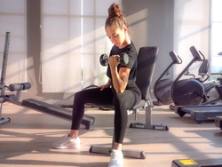 Beautiful athletic muscular woman pumps up the muscles by one arm lifts dumbbell exercise on bench in fitness gym. Young sport girl gains strong muscles physical by weight lifted in fitness studio.