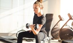 Sportive muscular woman pumps up the muscle by one arm lifts dumbbell exercise on bench in fitness gym. Young female athletic gain strong physical muscles by weight lifting workout in fitness studio.