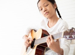 Pretty girl child and acoustic guitar isolated on white background. Kid guitarist put fingers on fingerboard of string instrument playing 5th fret songs. Selective focus on right-hand fingerpicking.