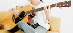 Young girl child and acoustic guitar sit on sofa white background. Kid guitarist put fingers on fingerboard of string instrument playing 3th fret songs. Selective focus on right-hand fingerpicking.