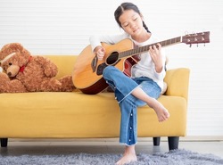 Talented child musician and acoustic guitar sit on couch white background. Female kid guitarist put fingers on fingerboard of string instrument playing fingerpicking melody. Pretty girl leisure hobby.