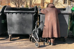 elderly woman in winter coat is looking for food in street garbage cans. disabled grandmother is rummaging for some food in garbage. poverty among the elderly