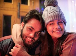 Happy Indian brother and sister sitting together at home in winters and embracing each other with a toothy smile and looking at the camera. They are wearing warm clothes, sweaters, jacket andcap.