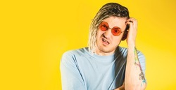 Young thoughtful handsome man in sunglasses, blue t-shirt scratching head. Pensive conceptual bristly guy with doubt glance, pondering facial expression on isolated yellow background in studio
