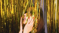 Young woman hiding face under golden tinsel in studio. Portrait of female with bright manicure on hands playing with hanging festive, sparkling decoration