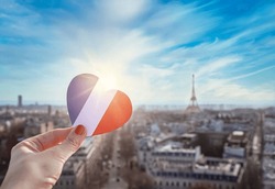 Girl's hand holding heart with french flag colors. Heart is on background of historic city center of Paris, famous Eiffel Tower, blue sky, clouds, sun. Love, Paris, France, travel concept.