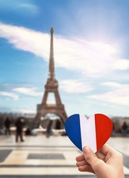 Girl's hand holding heart with french flag colors. Heart is on background of famous Eiffel Tower, blue sky, clouds, sun. Love, Paris, travel concept.