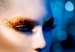 Beauty Fashion Model Girl with Holiday Leopard Makeup. Yellow Wild Cat Eyes Make-up Eyeshadow. Beautiful Woman Face with Perfect skin. Animal Make up. Blue Abstract Bokeh Background