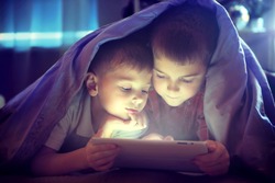 Two kids using tablet pc under blanket at night. Brothers with tablet computer in a dark room