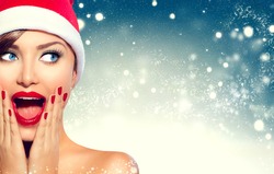 Christmas Girl. Beauty model woman in Santa Claus hat with red lips and manicure looking left with a surprised expression. Closeup portrait over winter snow wide background with copy space. Sales