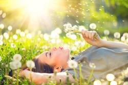 Beautiful Young Woman lying on the field in green grass and blowing dandelion. Outdoors. Enjoy Nature. Healthy Smiling Girl on spring lawn. Allergy free concept. Freedom