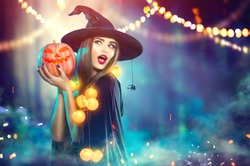 Halloween Witch with a carved Pumpkin and magic lights in a dark forest. Beautiful young surprised woman in witches hat and costume holding pumpkin. Halloween party art design