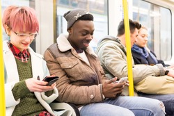 Multiracial smiling best friends sitting together in underground metro during commuting with mobile phones in their hands. Happy passengers in winter clothes use smartphones in public transportation.