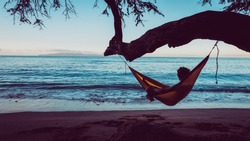 Traveler guy is relaxing in a hammock hang on tree directly on the beach. Man take a chilling moment during adventure vacation and enjoy the beauty of the Nature. Wanderlust and travel concept.