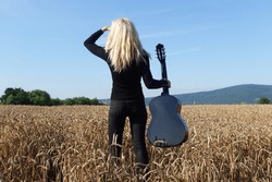 young woman posing in rye or wheat field. girl holding musical instrument called guitar and looking around. summer wallpaper. concert in nature.