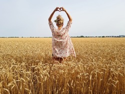 young woman posing in agriculture rye or barley or wheat field. girl wearing flower wreath on head and making heart shape with hands. nature lover. summer wallpaper.