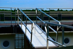 Sloped aluminum gangway or platform to board a ship. Pipe railing and chain. access bridge spanning across of river shore and boat. green forest and blue sky in the background.