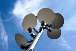Large white dish shaped light deflectors and reflector lamp heads attached  to vertical pole. blue sky and white clouds. low angle view. city lighting concept. abstract tilted view. dutch angle.