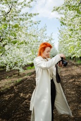 Girl photographer with red hair with a camera. female tourist, blogger, photographs nature. spring background. concept of work, hobby, tourism. spring light green tones.