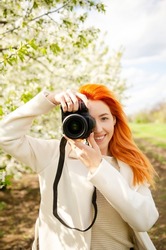 Girl photographer with red hair with a camera. female tourist, blogger, photographs nature. spring background. concept of work, hobby, tourism. spring light green tones.