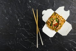 Chinese noodles in a box with chicken and vegetables with sticks. Wok food delivery from restaurant on dark background from top view.