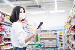 Asian woman wearing face mask and rubber glove push shopping cart in suppermarket departmentstore. Girl hold smartphone choose & look grocery things to buy during coronavirus crisis, covid19 outbreak.