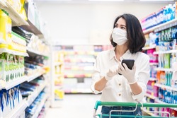 Asian woman wearing face mask and rubber glove push shopping cart in supermarket departmentstore. Girl hold smartphone choose & look grocery things to buy during coronavirus crisis, covid19 outbreak.