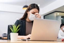 Sickness syndrome in office, Asian young businesswoman working on computer has headache, feeling stress and sick from work. Girl wearing mask preventing Covid19 infection from colleague in workplace.