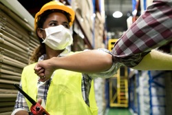 Two colleague female and male workers in warehouse factory have a greeting by touching the elbow or elbow bump to prevent the COVID infection. All people wear mask in industrial workplace for health.