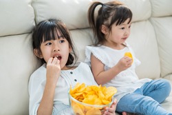 Asian Two sweet sisters little girl resting on sofa at home enjoy eating fast food, potato chips. Hungry preschool Cute kid Puts snack in mouth with hand. Tasty and unhealthy food for children concept