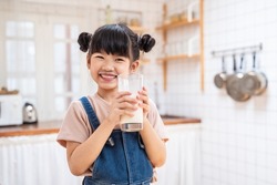 Portrait of Asian little cute kid holding a cup of milk in kitchen in house. Young preschool child girl or daughter stay home with smiling face, feel happy enjoy drinking milk and then look at camera.