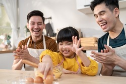 Asian LGBTQ male gay family teach girl kid stirring eggs in kitchen. Attractive handsome man couple take care and spend time with little adorable child bake bakery, enjoy parenting activity at home.