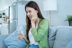 Asian beautiful woman feel terrible toothache after drink cold water. Attractive female sit on sofa in living room touching cheek, feel hurt and suffering from sensitive tooth ache, pain and cavities.