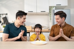 Asian attractive LGBTQ gay family celebrate birthday girl kid in house. Handsome male couple take care and surprise little adorable girl child with cake and present gift on her special day at home.