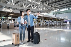 Asian man and woman passengers wear face mask walk in airport terminal. Young traveler couple stand in boarding gate during the COVID pandemic. New normal lifestyle in airport transportation concept.