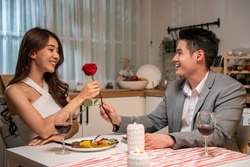 Asian young man surprise and give rose flower to beautiful girlfriend. Attractive romantic new marriage couple man and woman having dinner together to celebrate anniversary and valentines day in house