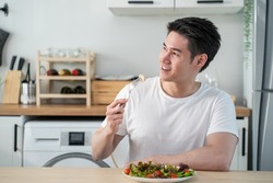 Asian attractive handsome male eating green salad in kitchen at home. Active young man sit on table, feel happy and enjoy vegetables healthy foods and clean water for breakfast to diet for health care