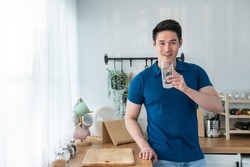 Portrait of Asian attractive male holding a glass of water in kitchen. Young thirsty handsome man pouring and drink clean mineral natural in cup after wake up in early morning for health care in house