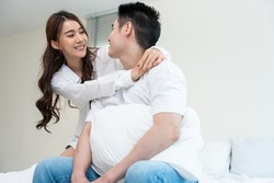 Asian new marriage couple sit on bed and look at each other with love. Attractive beautiful young man and woman in pajamas enjoy early morning activity in bedroom at home. Family relationship concept.
