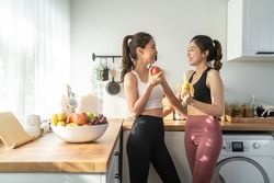 Asian active two women sibling in sportswear eat an apple in kitchen. Young beautiful girl sister feeling happy and enjoy eating fruits healthy foods to diet and lose weight for health care in house.