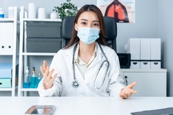 Asian young female medical doctor talking virtual online with patient. Attractive physician woman wear face mask, remote work from office hospital and broadcast diagnosis treatment to the sick people.